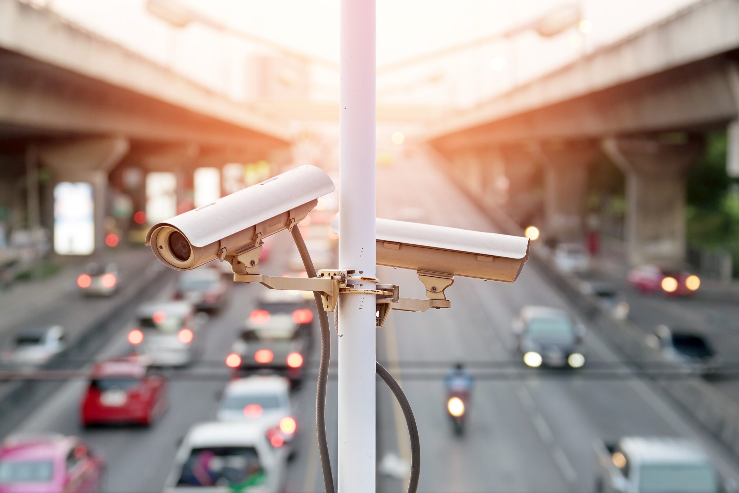 Traffic cameras on a lamppost with a background of cars on a busy main road.